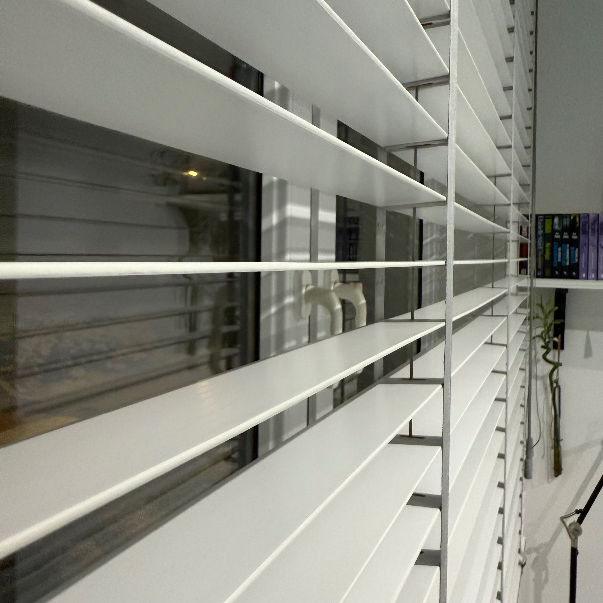 Białe żaluzje poziome zamontowane w oknie, widok z boku, z widocznym fragmentem wnętrza i półką z książkami w tle. Delikatne światło podkreśla strukturę żaluzji.