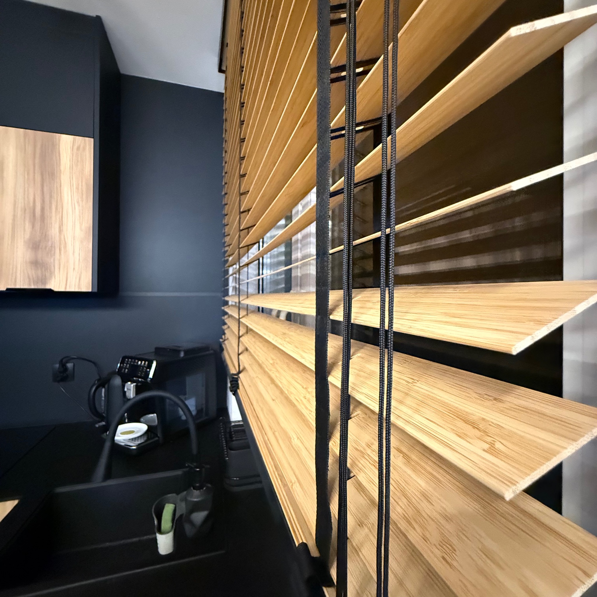 Zamknięte żaluzje drewniane w odcieniach beżu, z czarnymi sznurkami, na tle ciemnej kuchni z czarnym zlewem i ekspresem do kawy. Ujęcie z dołu, podkreślające strukturę drewna.