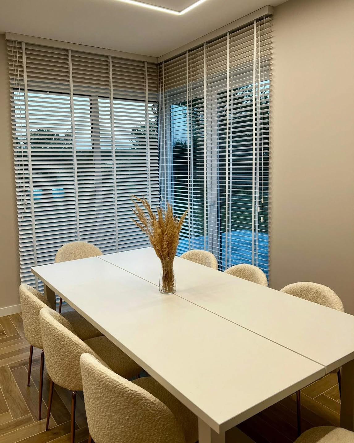 Jasne drewniane żaluzje poziome w nowoczesnej jadalni z białym stołem i beżowymi krzesłami. Dekoracyjny bukiet w wazonie na stole. Minimalistyczny design.