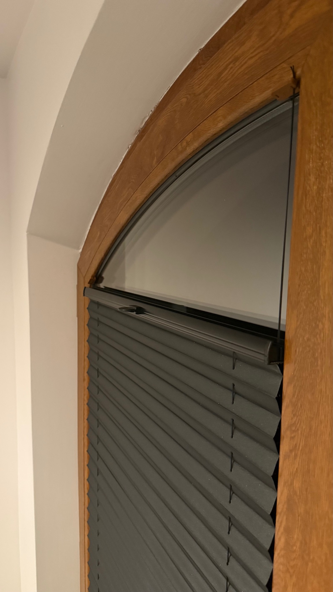 Szara plisa okienna zamontowana w drewnianej, łukowej ramie okiennej na tle białej ściany. Widoczne detale konstrukcji i faktura materiału.