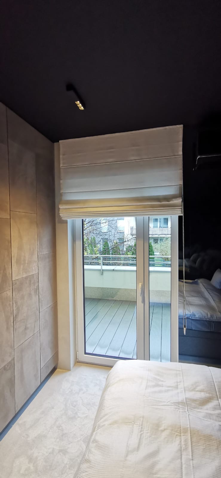 Elegancka roleta rzymska w jasnym odcieniu, zamontowana nad oknem balkonowym w nowoczesnej sypialni z ciemnym sufitem i jasną podłogą. Widok na balkon.