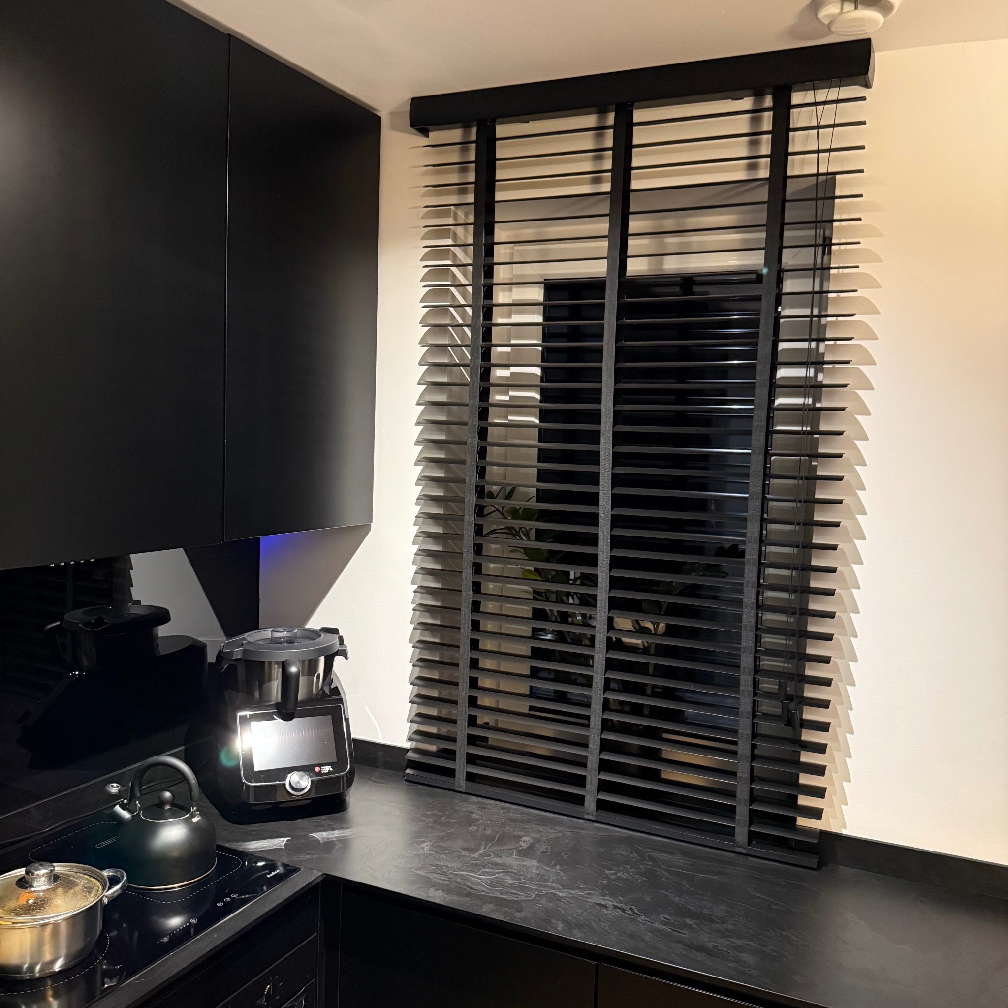 Czarne żaluzje drewniane w nowoczesnej kuchni z czarnymi meblami i sprzętami. Widok na okno z rośliną w tle. Minimalistyczny design i eleganckie wykończenie.