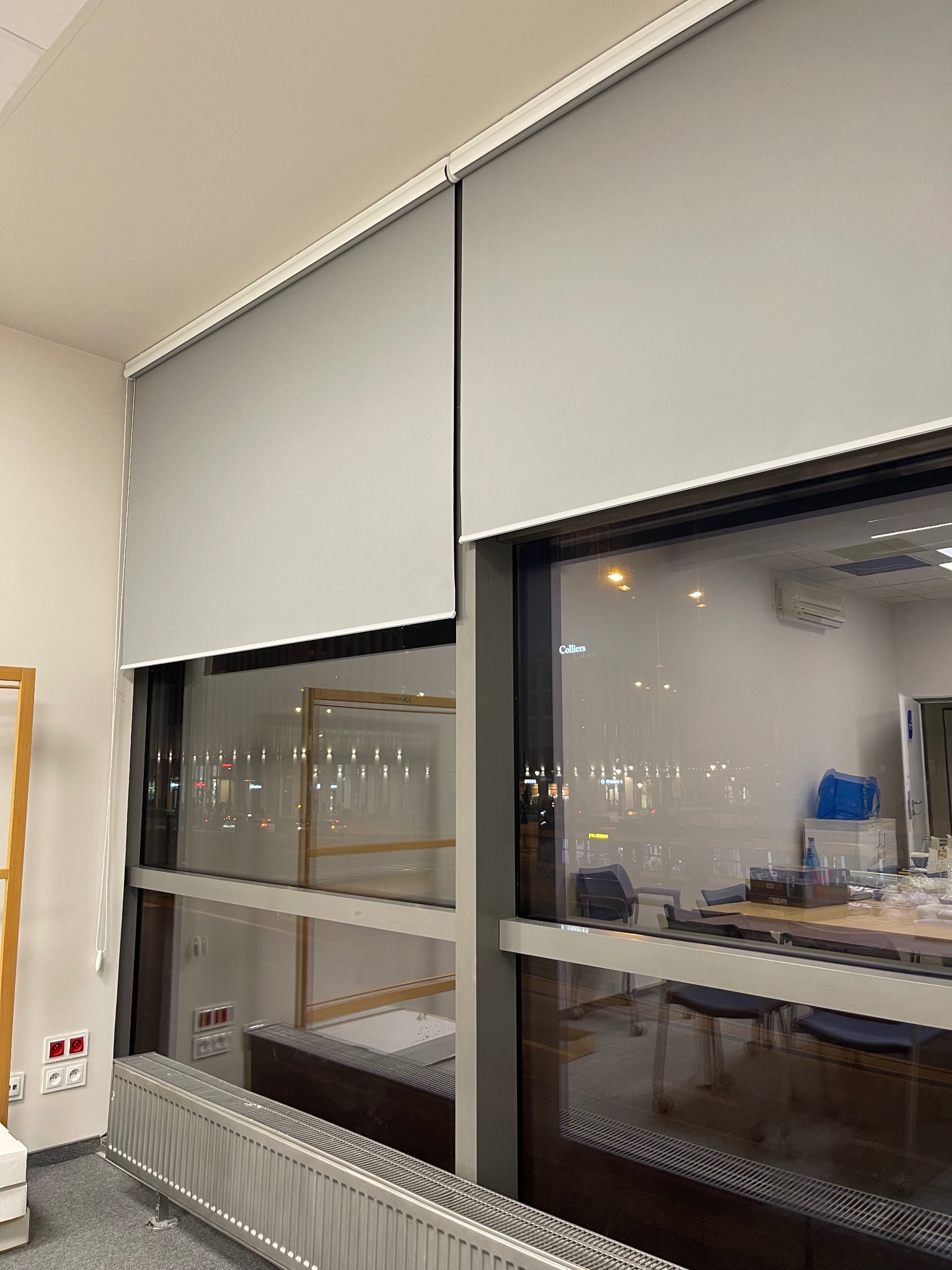 Dwie szare rolety wewnętrzne zamontowane w oknach biurowych. Widoczny mechanizm rolety i fragment wnętrza biura z widokiem na miasto nocą.