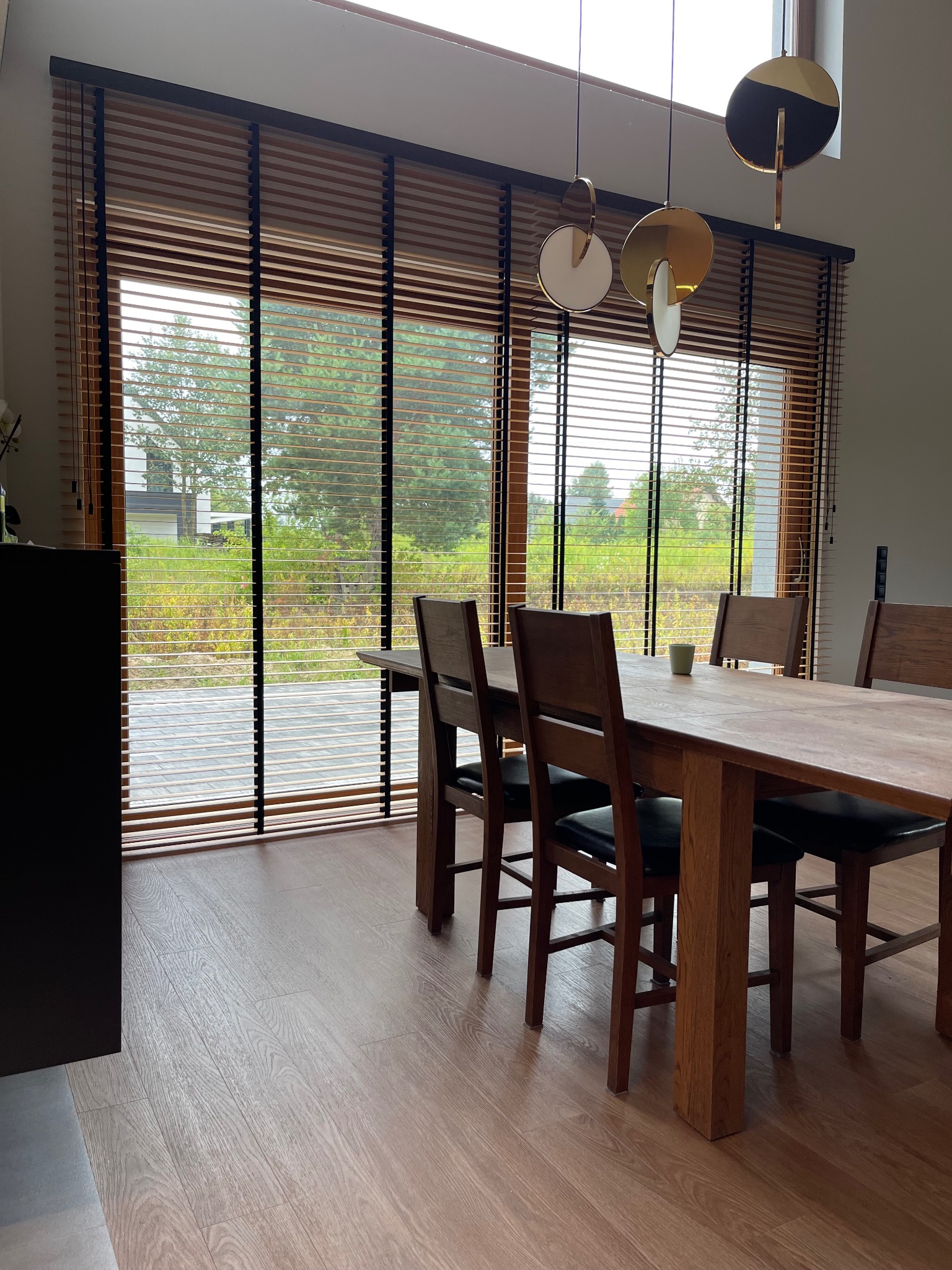Drewniane żaluzje poziome w Łodzi, zamontowane w oknie jadalni, z widokiem na zieleń. Nowoczesne lampy wiszące i drewniany stół dodają wnętrzu ciepła.