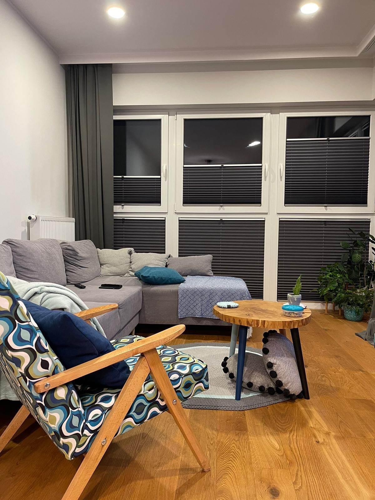 Ciemne plisy okienne w nowoczesnym salonie z szarą sofą i drewnianym stolikiem kawowym. Widok na okna z plisami w neutralnym wnętrzu.