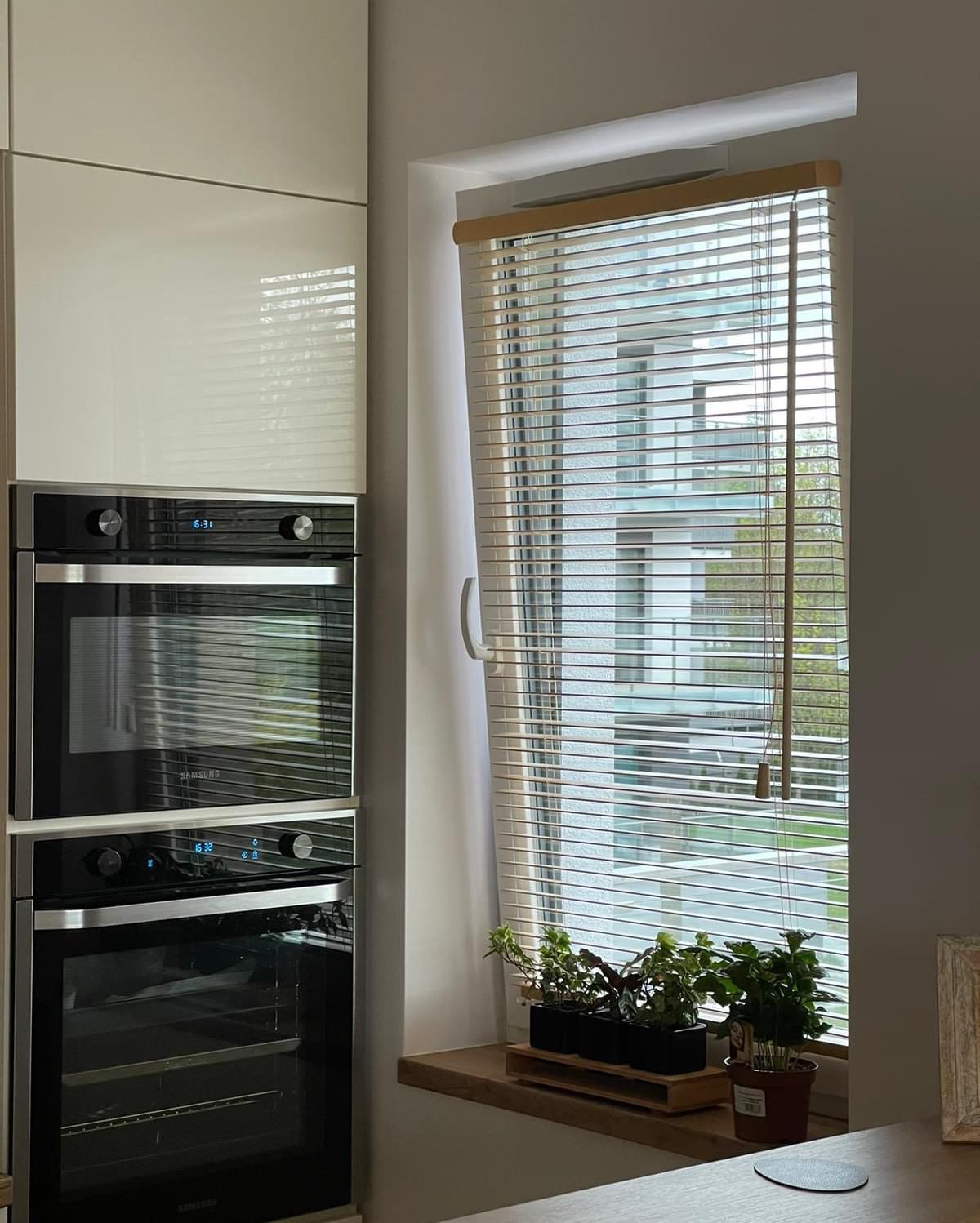 Jasne żaluzje poziome z drewnianą listwą górną, zamontowane w oknie. Na parapecie doniczki z roślinami. W tle zabudowa kuchenna z piekarnikami.
