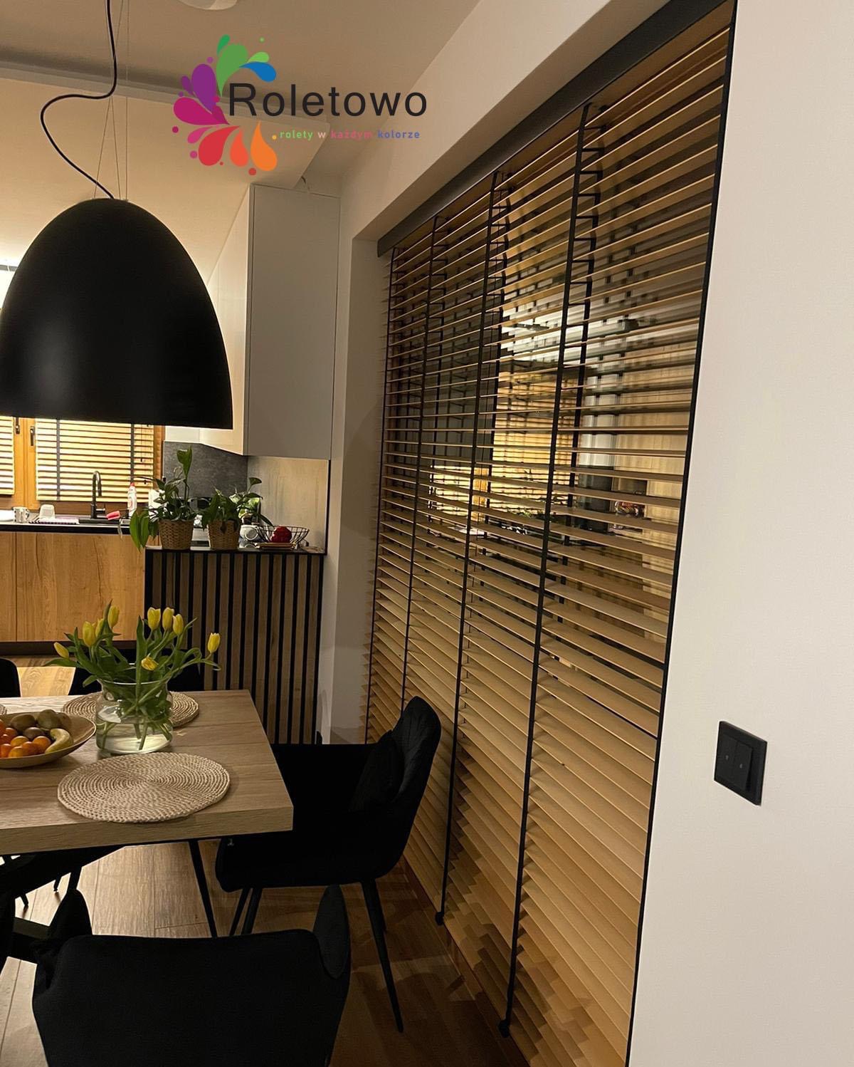 Jasne żaluzje z ciemnymi listwami w nowoczesnym wnętrzu. Widok na stół z tulipanami i czarne krzesła. Minimalistyczny design, ciepłe światło.