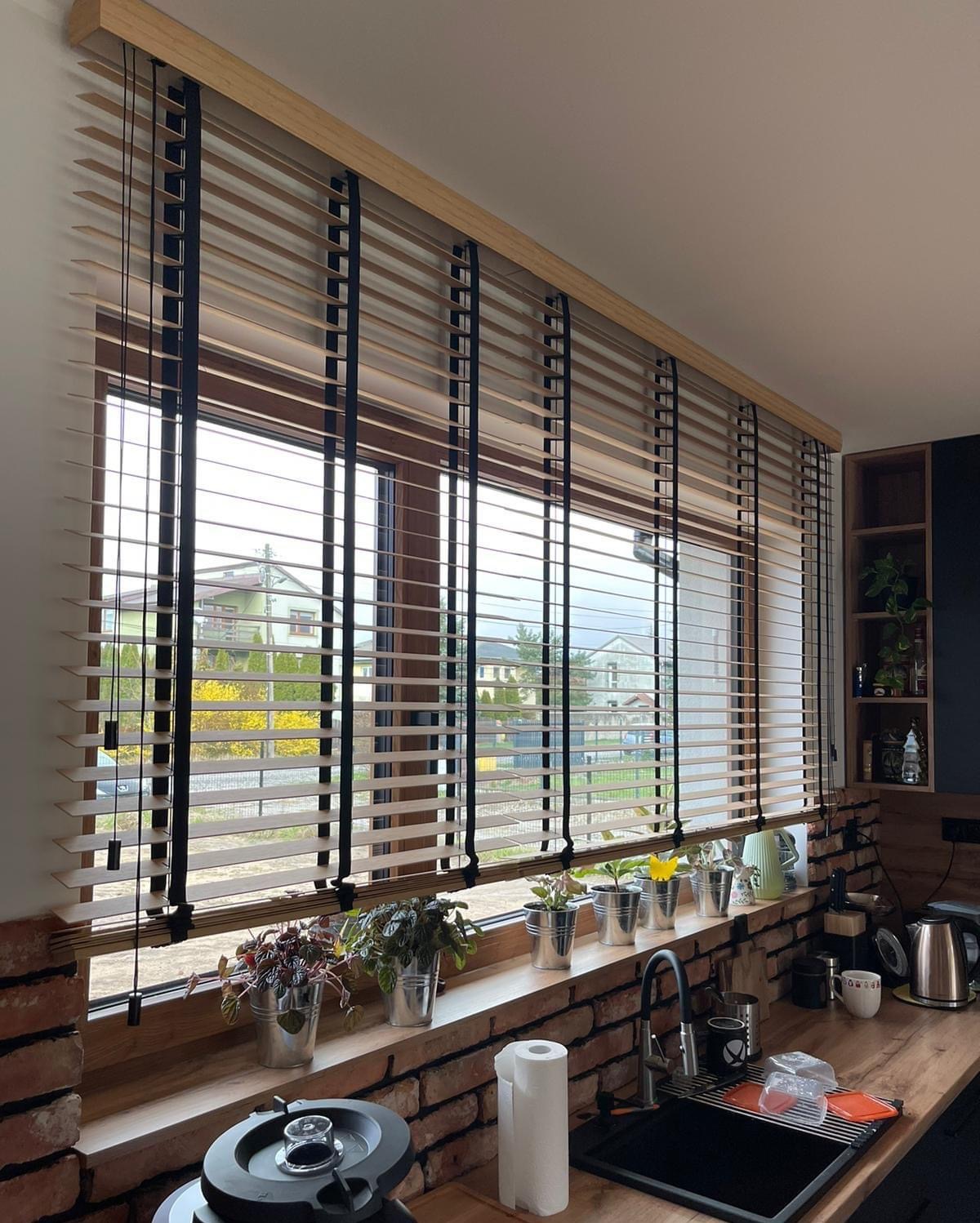 Drewniane żaluzje poziome z czarnymi taśmami w oknie kuchni z ceglanym blatem i ziołami w doniczkach. Widok na domy za oknem.