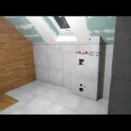 Łazienka w trakcie wykańczania: szare płytki na ścianach, drewnopodobne na skosie, sufit z płyt karton-gips, okno dachowe. Widoczne puszki elektryczne i instalacja.