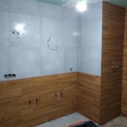 Agnieszka Gołębiewska - Łazienka w trakcie wykańczania: częściowo położone płytki imitujące drewno na ścianach, sufit z płyt karton-gips, widoczne instalacje i narzędzia glazurnicze.