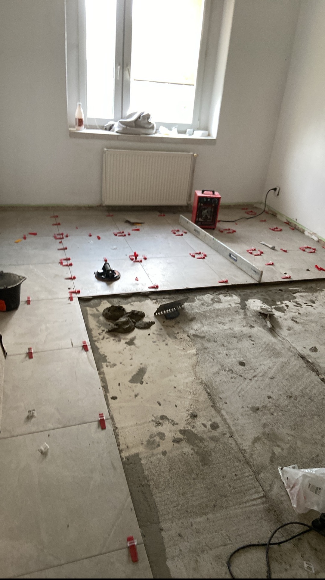 Układanie jasnych płytek podłogowych w pokoju, z użyciem poziomicy i systemu poziomowania płytek. Częściowo położone płytki, widoczne narzędzia i zaprawa.