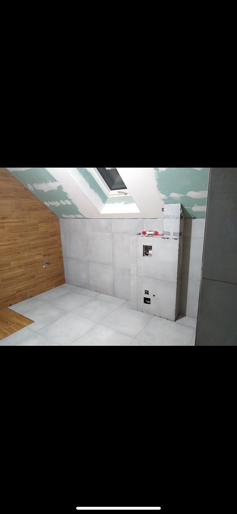 Łazienka w trakcie wykańczania: szare płytki na ścianach, drewnopodobne na skosie, sufit z płyt karton-gips, okno dachowe. Widoczne puszki elektryczne i instalacja.