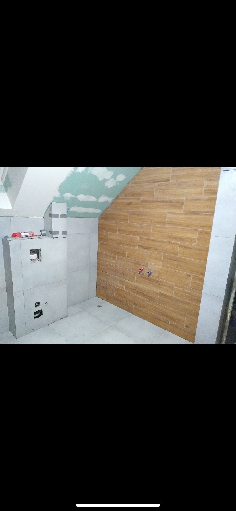 Łazienka w trakcie remontu: drewnopodobne płytki na ścianie, sufit z płyt karton-gips, szara zabudowa WC. Instalacja wodna widoczna, podłoga wyłożona jasnymi płytkami.