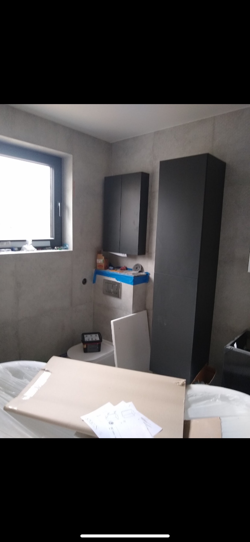 Łazienka w trakcie montażu: szare płytki, wiszące szafki, częściowo rozpakowana wanna i kartony z instrukcjami. Widoczne narzędzia i zabezpieczenia na ścianie.