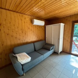 Air Balance Hubert Górka - Wnętrze pokoju z drewnianą boazerią, szarą sofą i klimatyzatorem ściennym. Widoczna biała szafa i drzwi balkonowe z widokiem na zieleń.