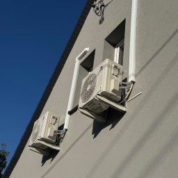 Air Balance Hubert Górka - Dwa klimatyzatory marki Gree zamontowane na szarej ścianie budynku z widocznymi przewodami i rurami odprowadzającymi, na tle błękitnego nieba.