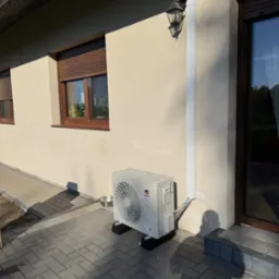 Zamontowana jednostka zewnętrzna klimatyzacji Gree na elewacji domu z brązowymi oknami i kostką brukową. Widoczna lampa zewnętrzna i maskownica instalacji.