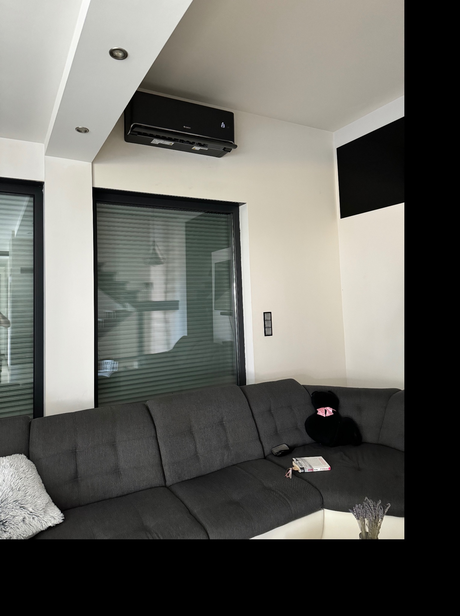 Czarny klimatyzator ścienny zamontowany w nowoczesnym wnętrzu nad szarą sofą, widoczne okno z żaluzjami i elementy dekoracyjne.