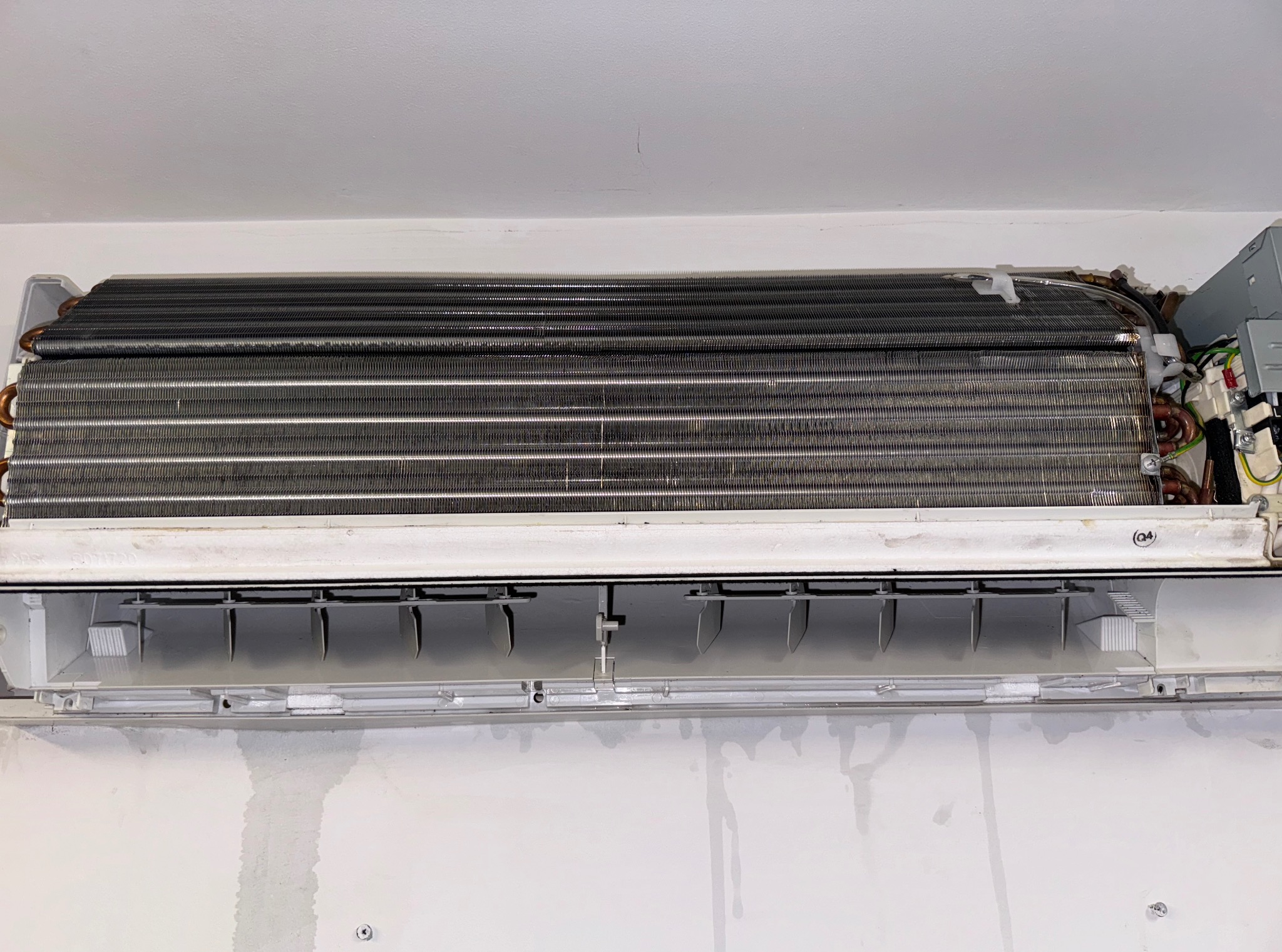 Otwarta, biała klimatyzacja ścienna ukazuje metalowe żebra chłodzące i elementy elektryczne. Ściana pod urządzeniem ma ślady po czyszczeniu.