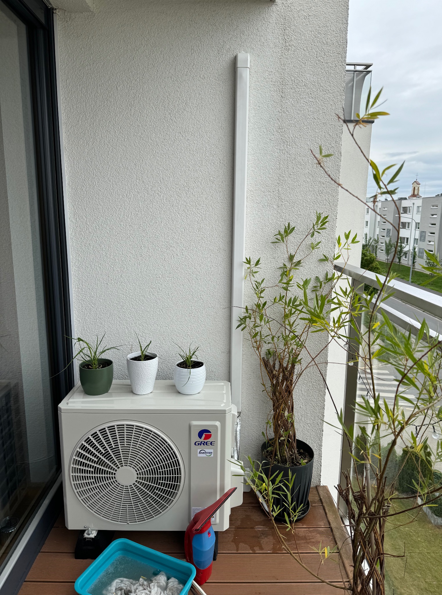 Kompaktowa jednostka klimatyzacji Gree na balkonie z drewnianą podłogą, otoczona roślinami w doniczkach. Widoczne narzędzia czyszczące i fragment zabudowy miejskiej w tle.