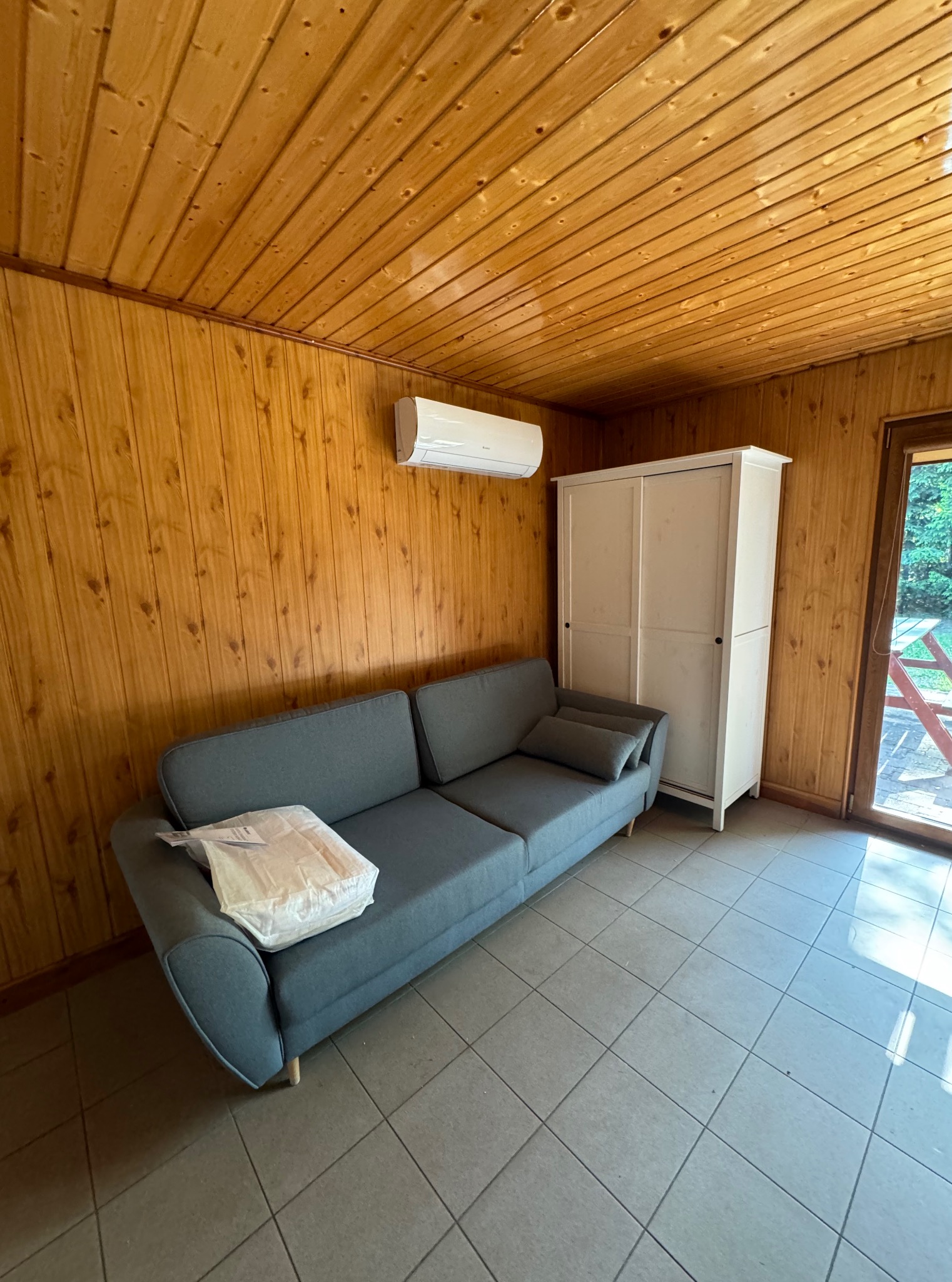 Wnętrze pokoju z drewnianą boazerią, szarą sofą i klimatyzatorem ściennym. Widoczna biała szafa i drzwi balkonowe z widokiem na zieleń.