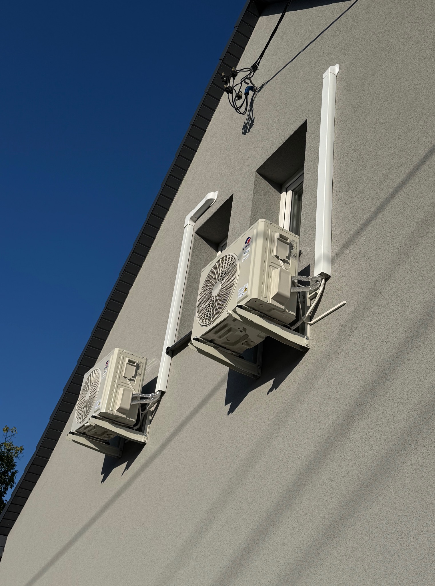 Dwa klimatyzatory marki Gree zamontowane na szarej ścianie budynku z widocznymi przewodami i rurami odprowadzającymi, na tle błękitnego nieba.