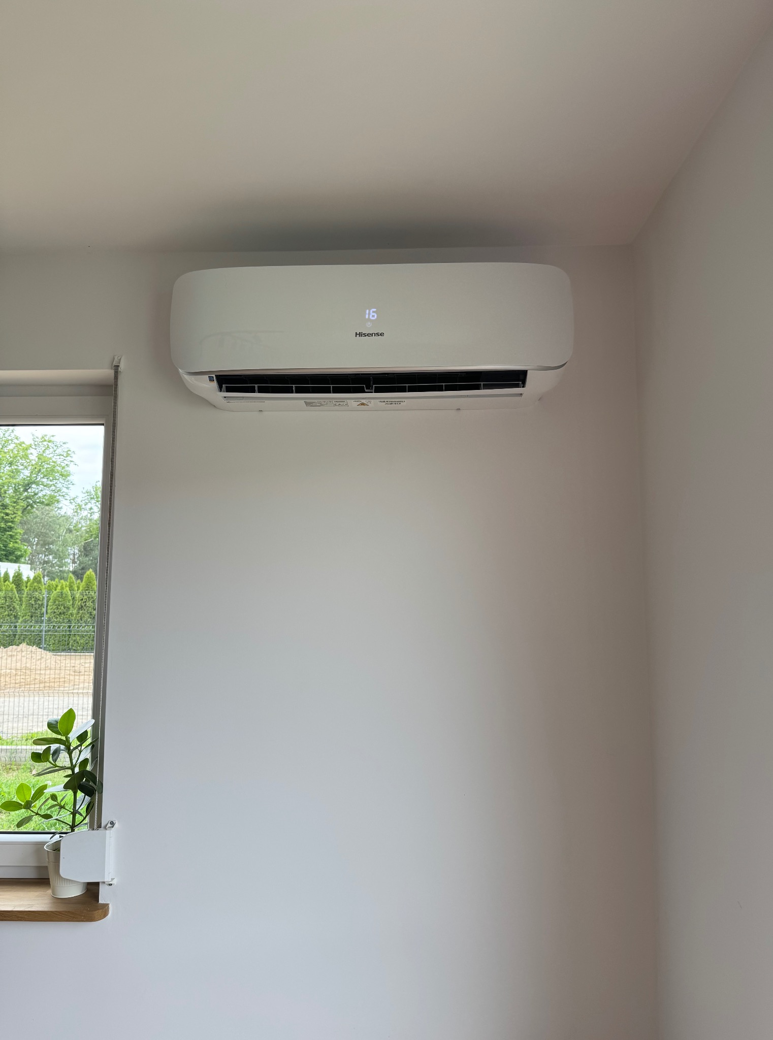 Biała klimatyzacja Hisense zamontowana na białej ścianie w jasnym pomieszczeniu, obok okna z rośliną. Widoczny wyświetlacz z temperaturą 16 stopni.