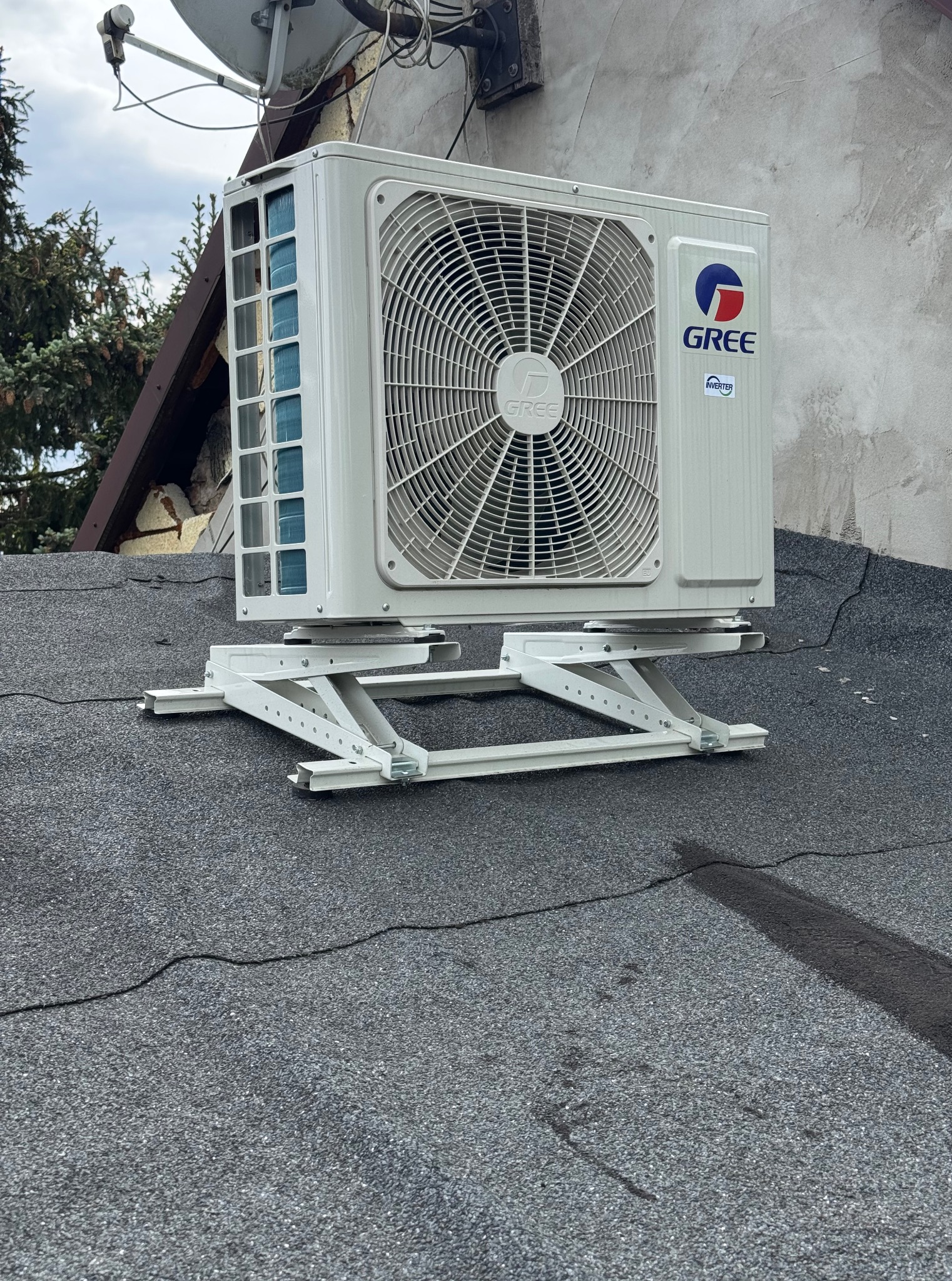 Biała jednostka zewnętrzna klimatyzacji marki Gree zamontowana na dachu budynku, na specjalnej, metalowej konstrukcji wsporczej. Widoczny fragment dachu pokrytego papą.