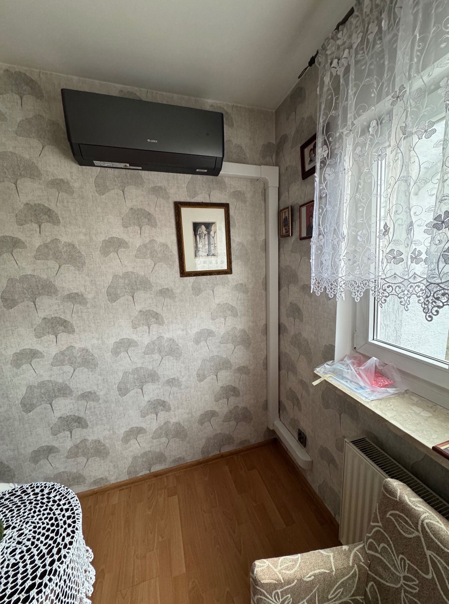 Szary klimatyzator marki Gree zamontowany na ścianie w pokoju z tapetą w liście. Widoczne rury instalacyjne, okno z firanką i fragmenty mebli.