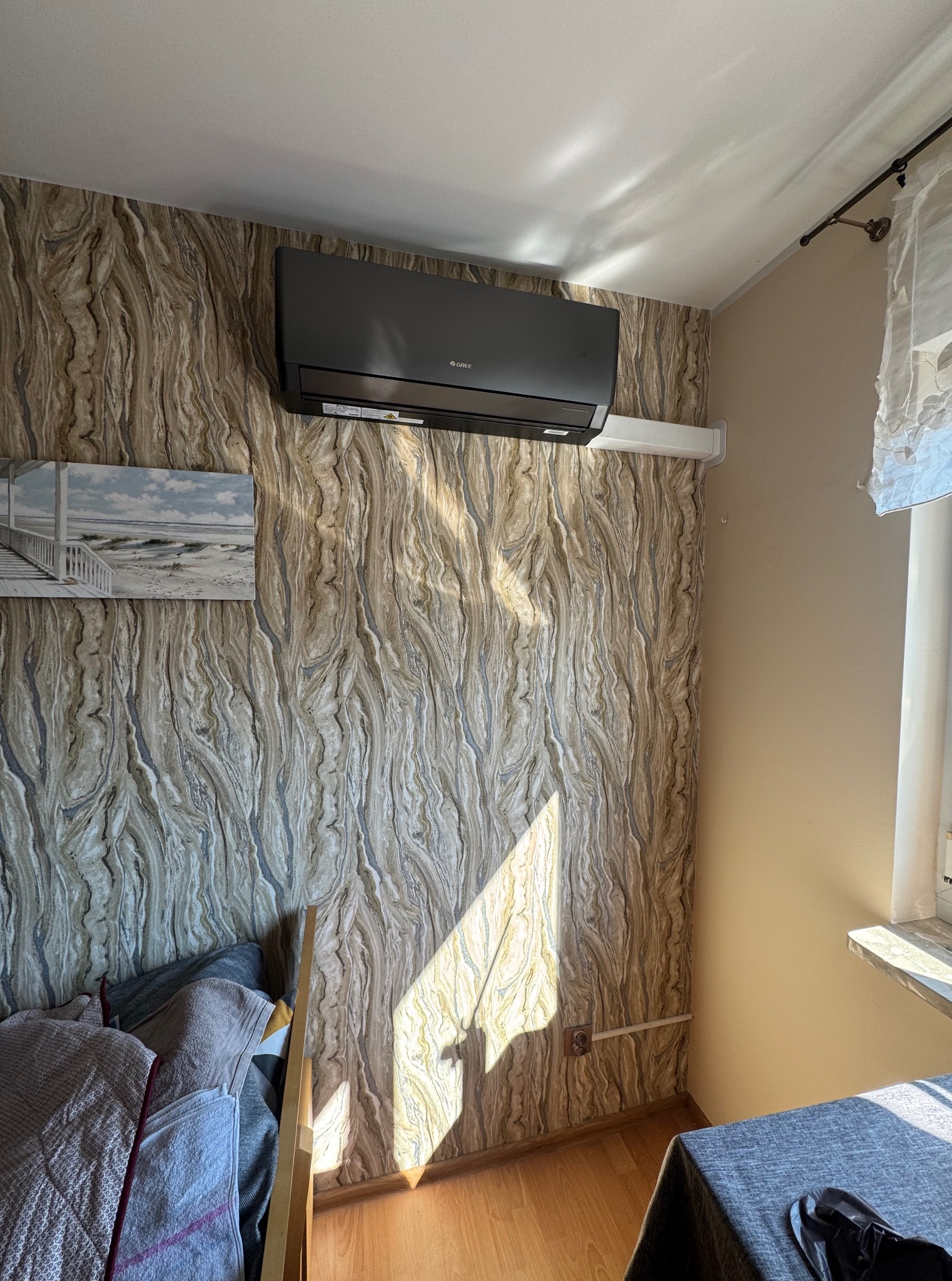 Czarny klimatyzator Gree zamontowany na wzorzystej ścianie w pokoju. Widoczny fragment łóżka i obrazek z motywem morskim. Promień słońca oświetla ścianę.