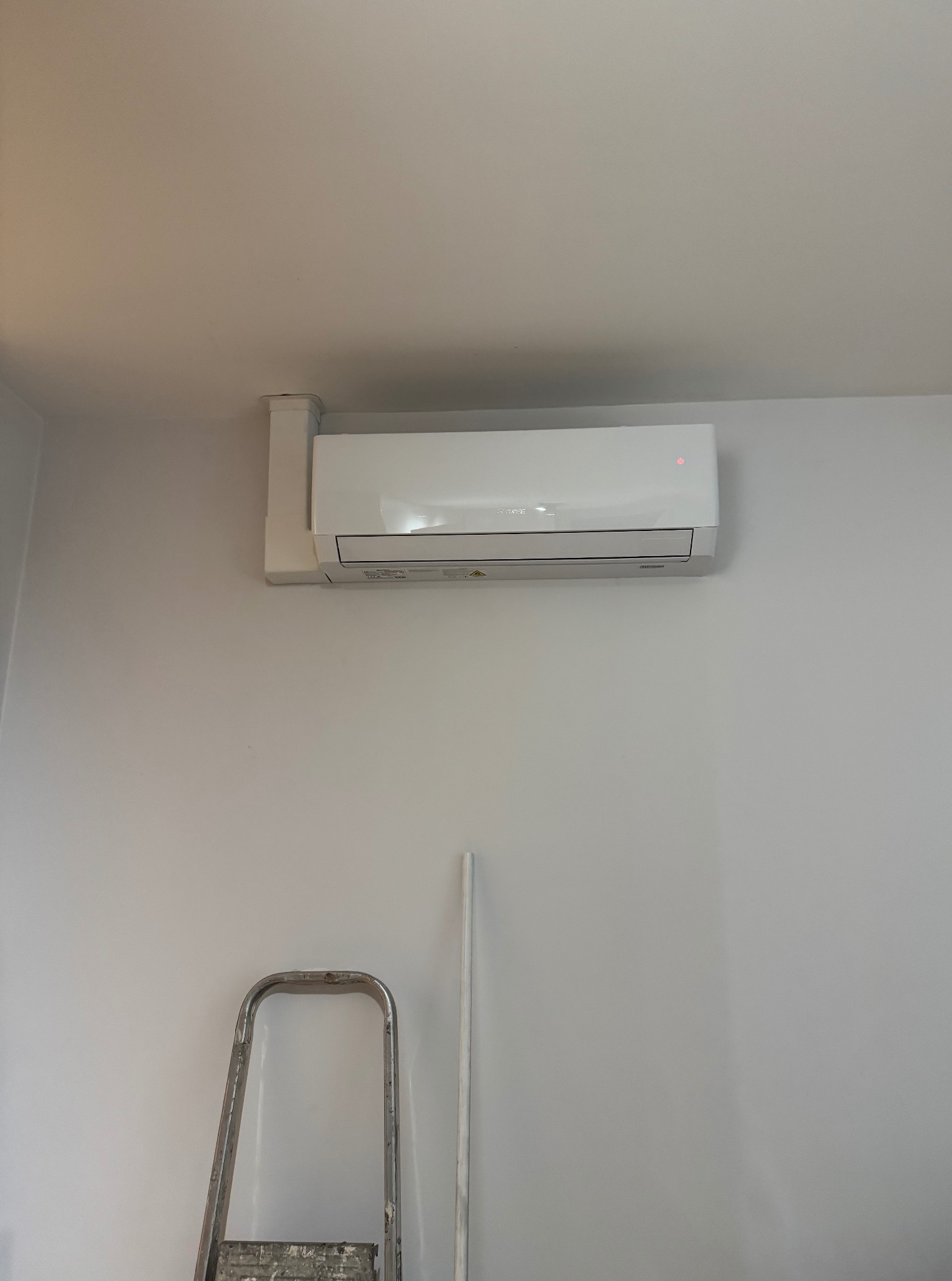 Biała klimatyzacja typu split zamontowana na ścianie, z widoczną instalacją i drabiną. Minimalistyczny design wnętrza, jasne ściany, proste linie urządzenia.