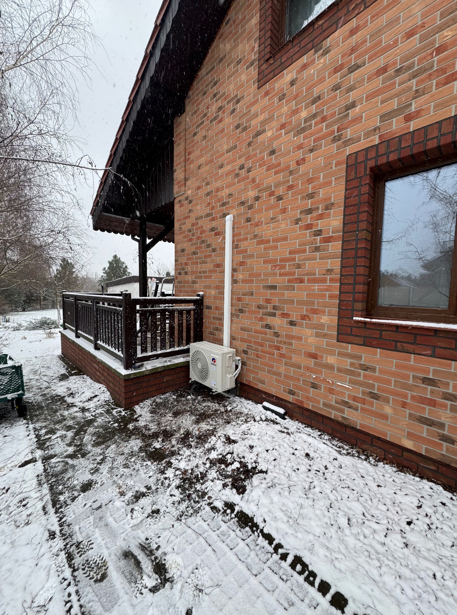 Zewnętrzna jednostka klimatyzacji na tle ceglanego domu w zimowej scenerii. Biała rura odprowadzająca skropliny. Śnieg na ziemi i werandzie.