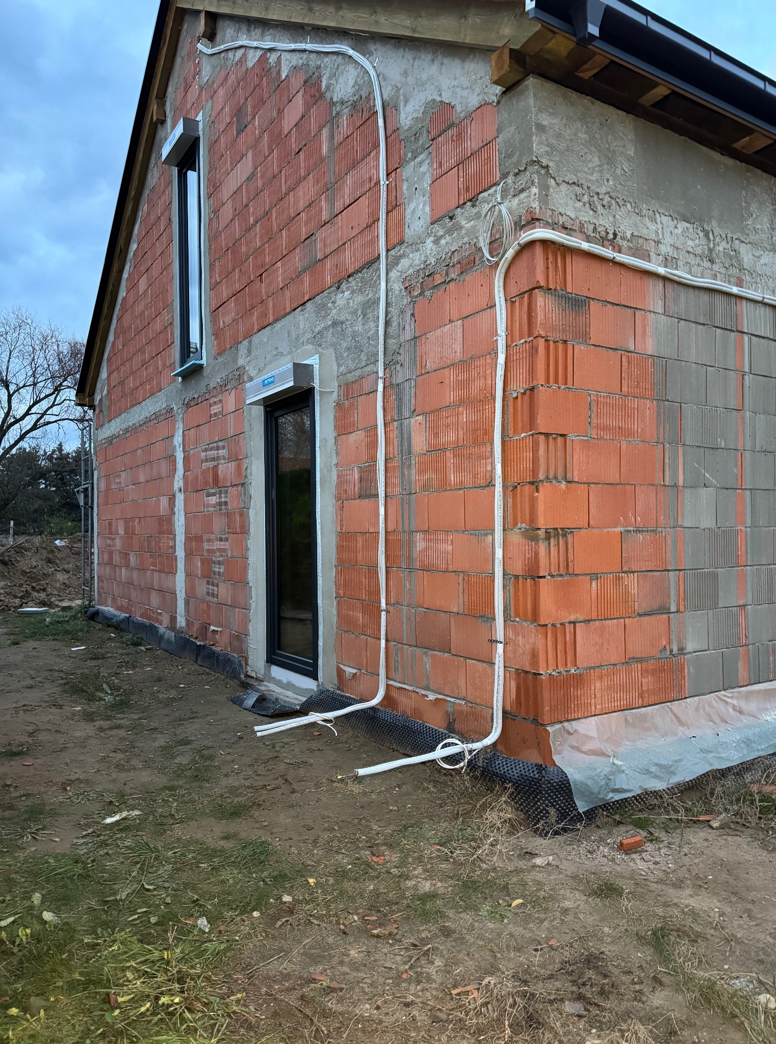 Surowy stan budynku z czerwonej cegły, z widoczną białą instalacją elektryczną biegnącą po elewacji, okna z roletami zewnętrznymi, teren budowy.