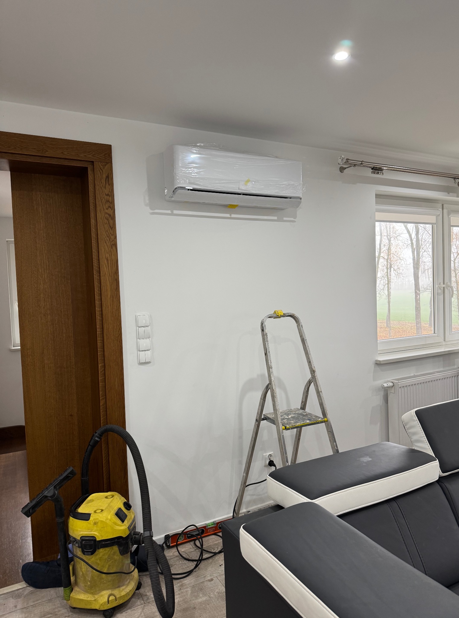 Świeżo zamontowany klimatyzator, jeszcze w folii ochronnej, na białej ścianie w salonie. Obok drabina i odkurzacz, sugerujące prace montażowe.