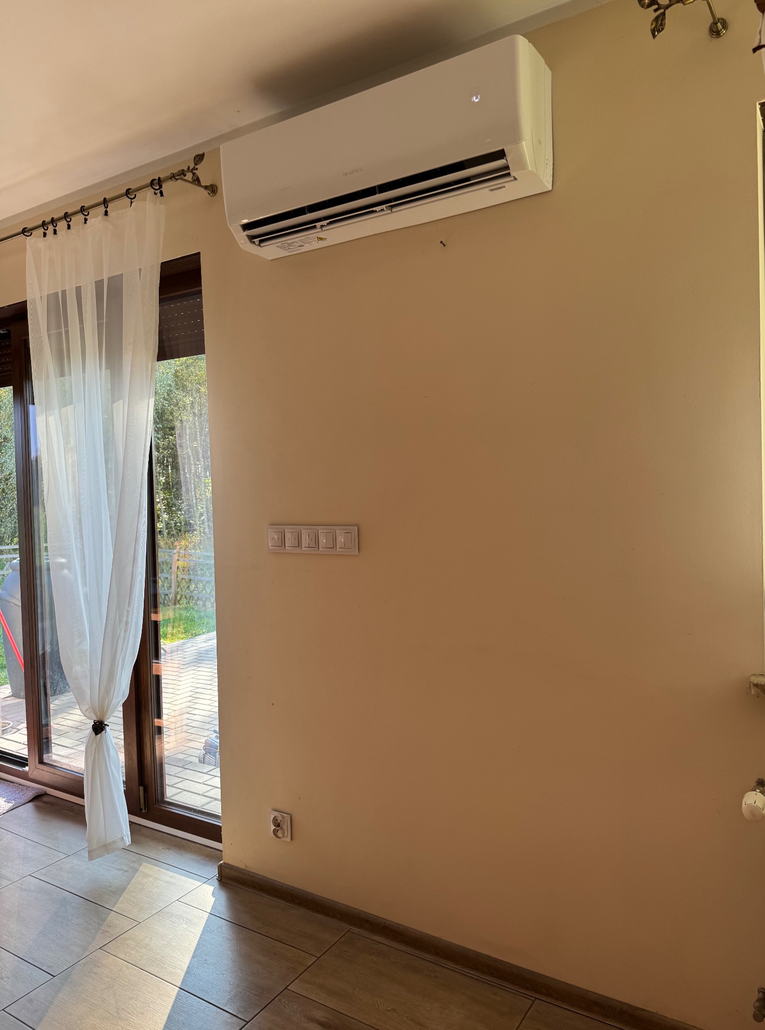 Zamontowana klimatyzacja na ścianie w jasnym pokoju z oknem balkonowym. Widoczne detale montażu i instalacja elektryczna. Świeże, jasne wnętrze.