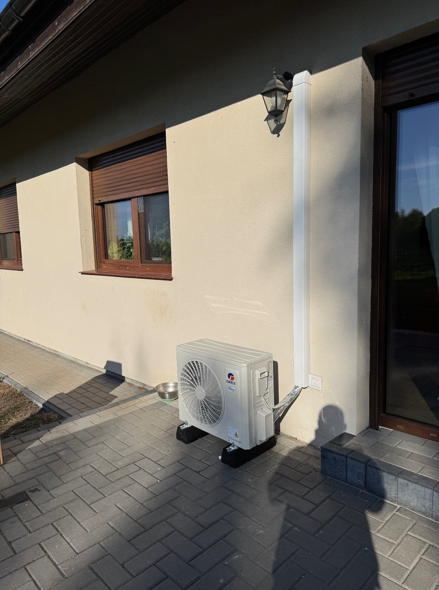Zamontowana jednostka zewnętrzna klimatyzacji Gree na elewacji domu z brązowymi oknami i kostką brukową. Widoczna lampa zewnętrzna i maskownica instalacji.