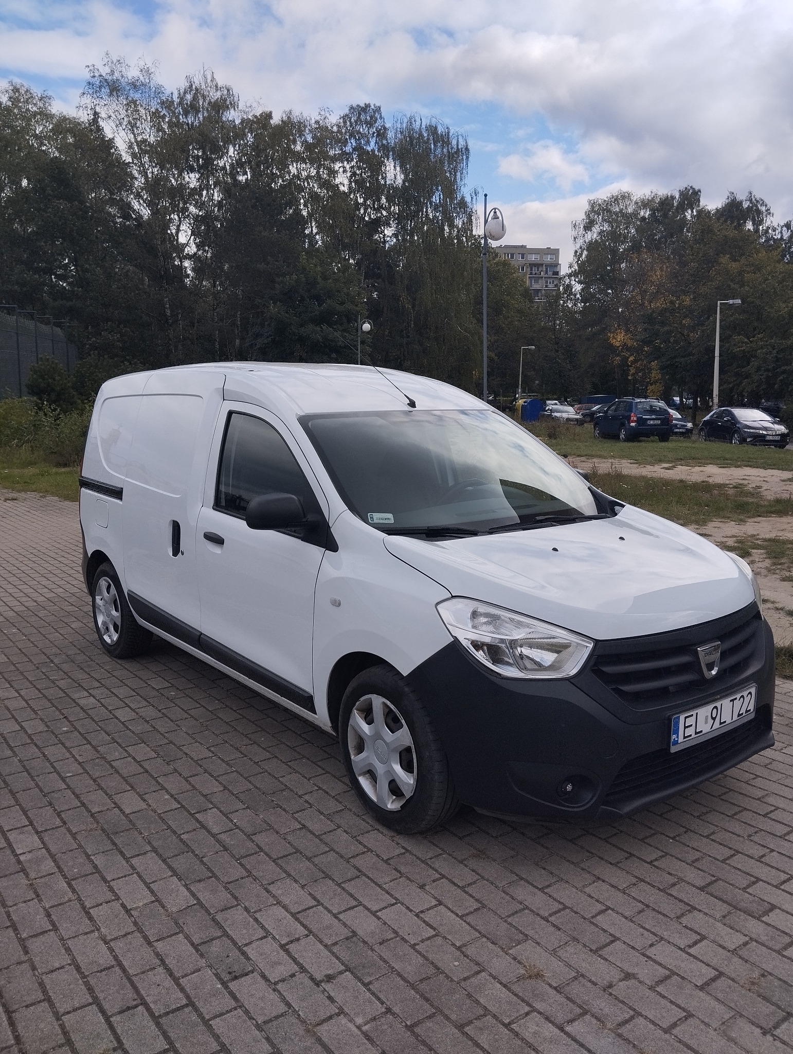 Biały bus Dacia Dokker Van na brukowanym placu, widok z boku, tablica rejestracyjna EL 9LT22, drzewa i budynek w tle.