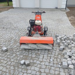 Mar Speed - Układanie kostki granitowej przy użyciu maszyny czyszczącej Boels. Widoczne kostki granitowe, garaż w tle. Etap prac wykończeniowych.