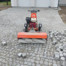 Układanie kostki granitowej przy użyciu maszyny czyszczącej Boels. Widoczne kostki granitowe, garaż w tle. Etap prac wykończeniowych.