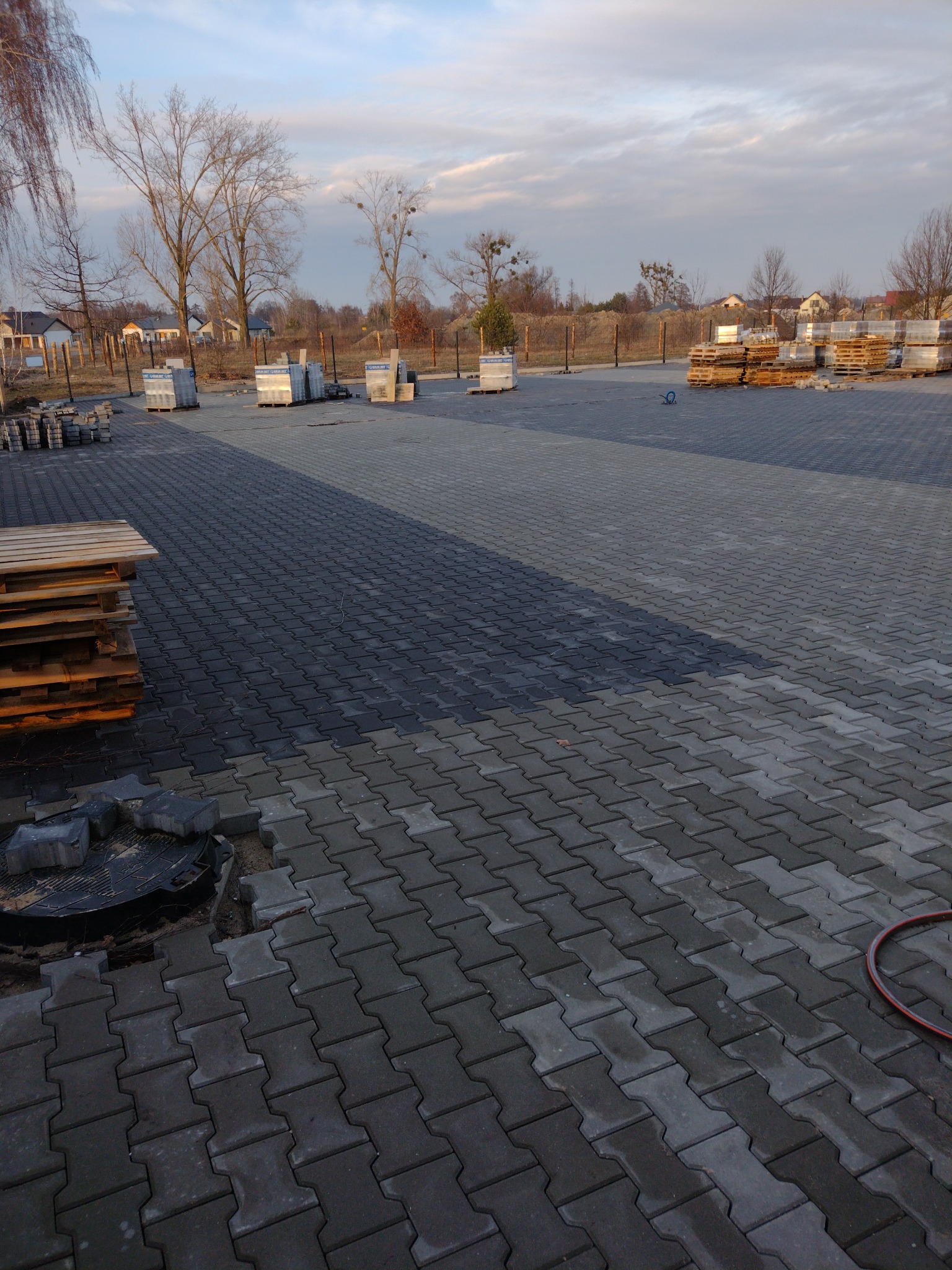 Ułożona szara kostka brukowa na dużej powierzchni, z paletami kostki w tle i nieukończonym fragmentem przy studzience kanalizacyjnej. Widoczne drzewa i zabudowania w oddali.