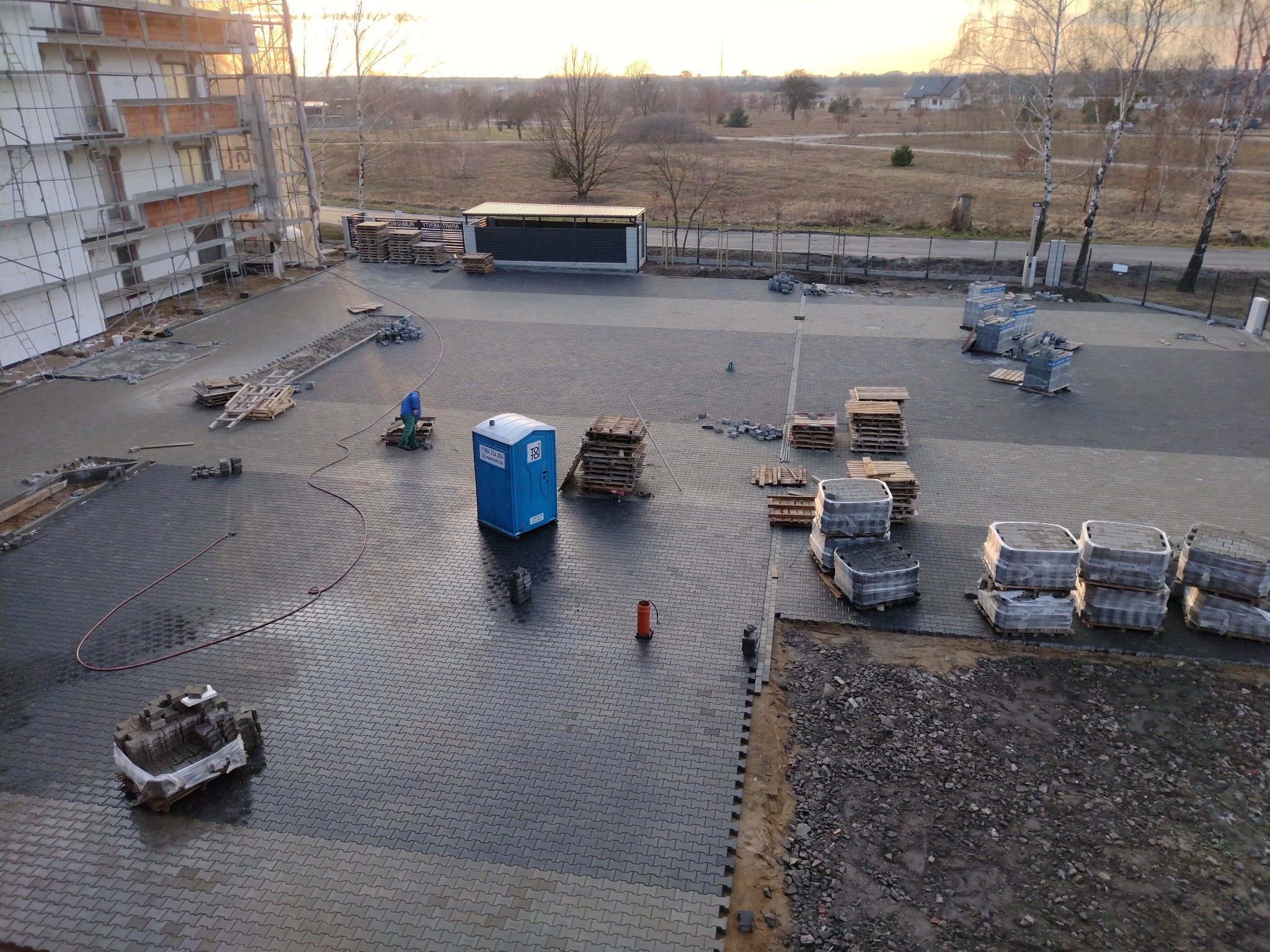 Szeroki kadr z góry na plac budowy z ułożoną szarą kostką brukową, paletami kostki, toaletą przenośną i fragmentem budynku w rusztowaniach. Widoczne drzewa w tle.