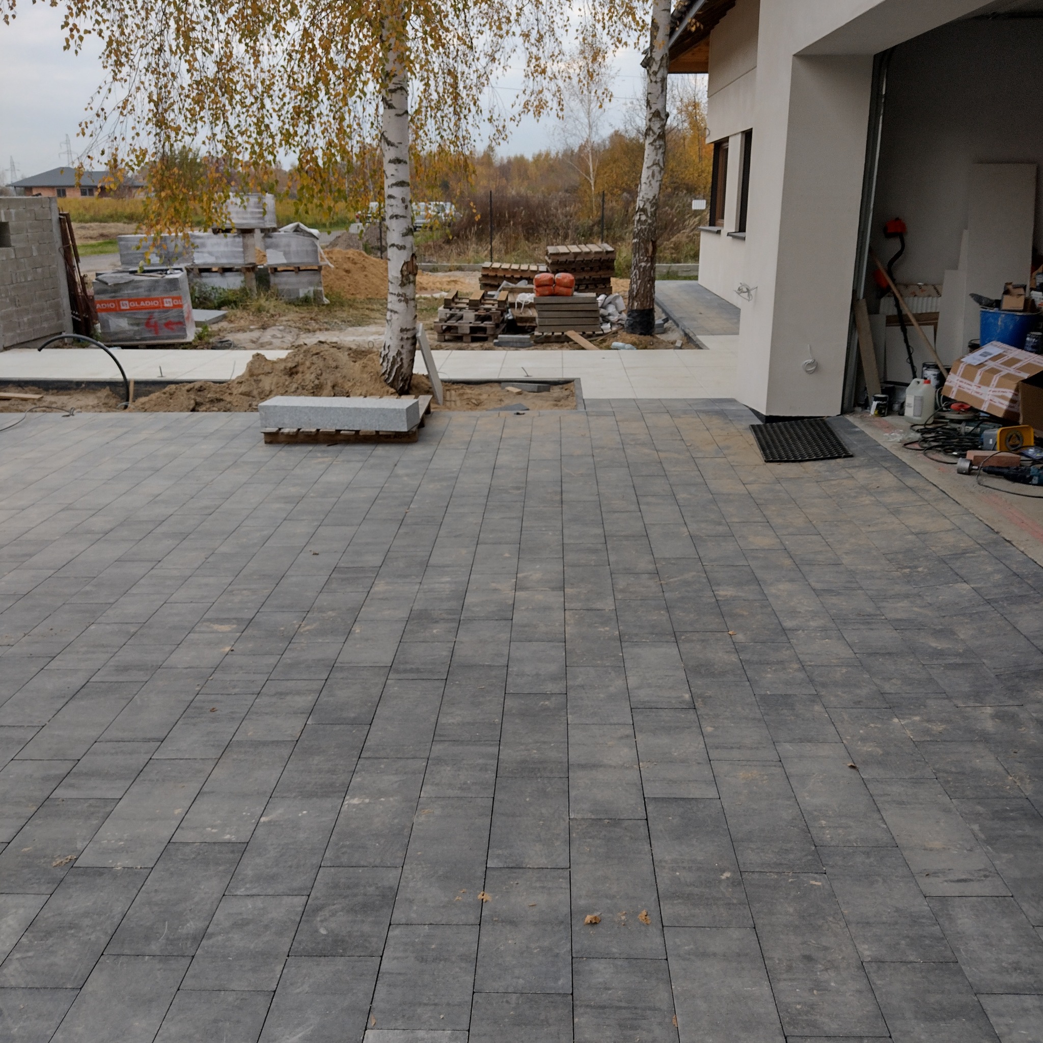 Ułożona, szara kostka brukowa na podjeździe domu z dwoma młodymi drzewami. Częściowo widoczne materiały budowlane i narzędzia. Jesienna sceneria w tle.