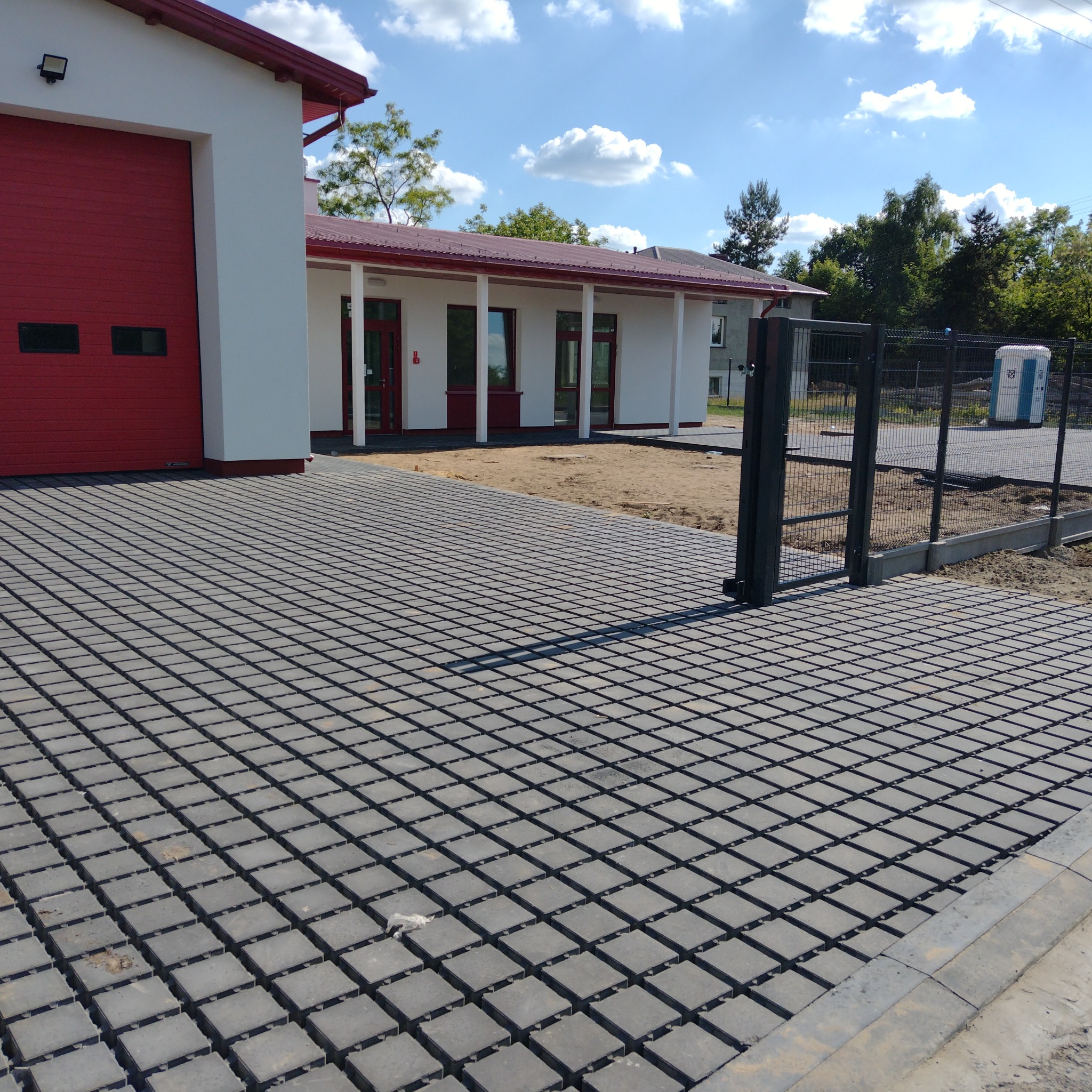 Szara ażurowa kostka brukowa przed budynkiem z czerwoną stolarką i szarą bramą wjazdową. Widoczne niebo z chmurami i zieleń w tle.