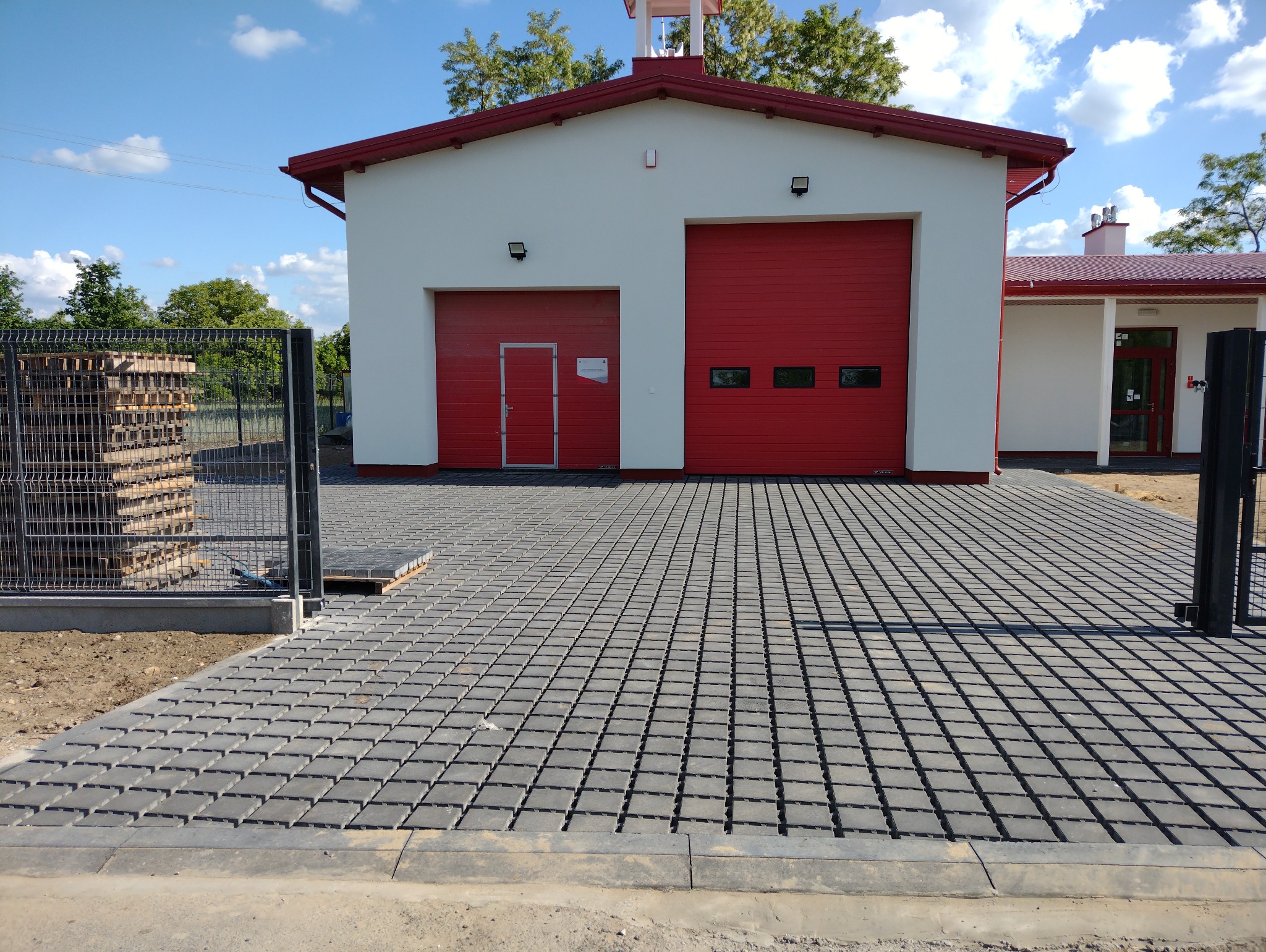 Szara, ażurowa kostka brukowa przed budynkiem remizy strażackiej z czerwoną bramą garażową i ogrodzeniem z paletami w tle, w słoneczny dzień.