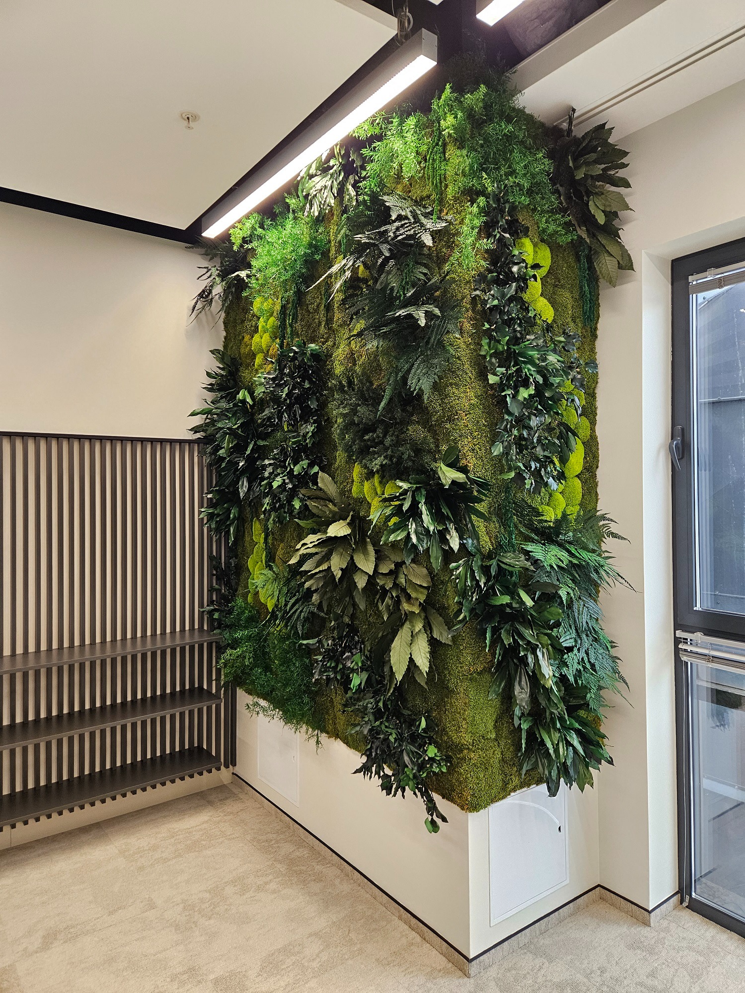 Wertykalny ogród z mchu i roślin w nowoczesnym wnętrzu biurowym, oświetlony lampą LED, obok okno i regał z pionowymi listwami. Dekoracja ściany.