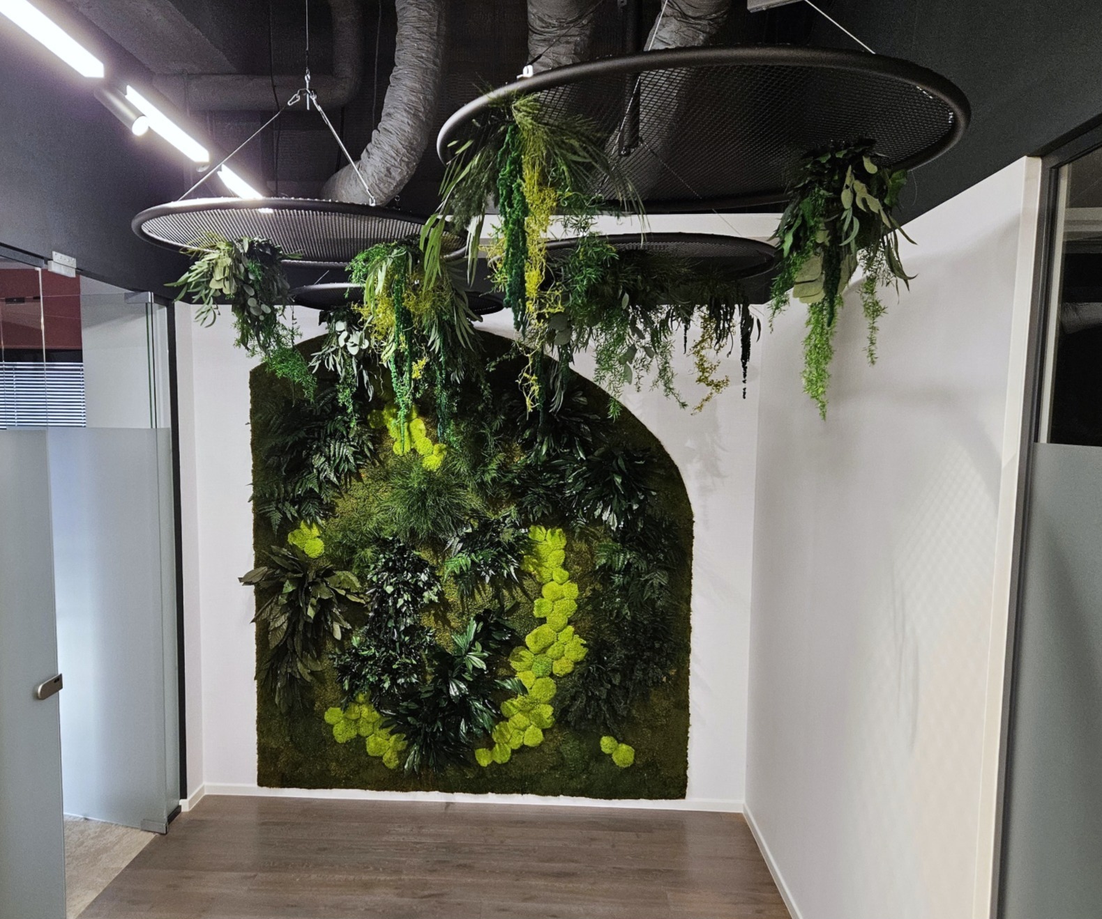 Wnętrze biura z zieloną ścianą z mchu i roślin, oświetloną przez okrągłe lampy sufitowe z wiszącymi pnączami. Nowoczesny design z elementami natury.