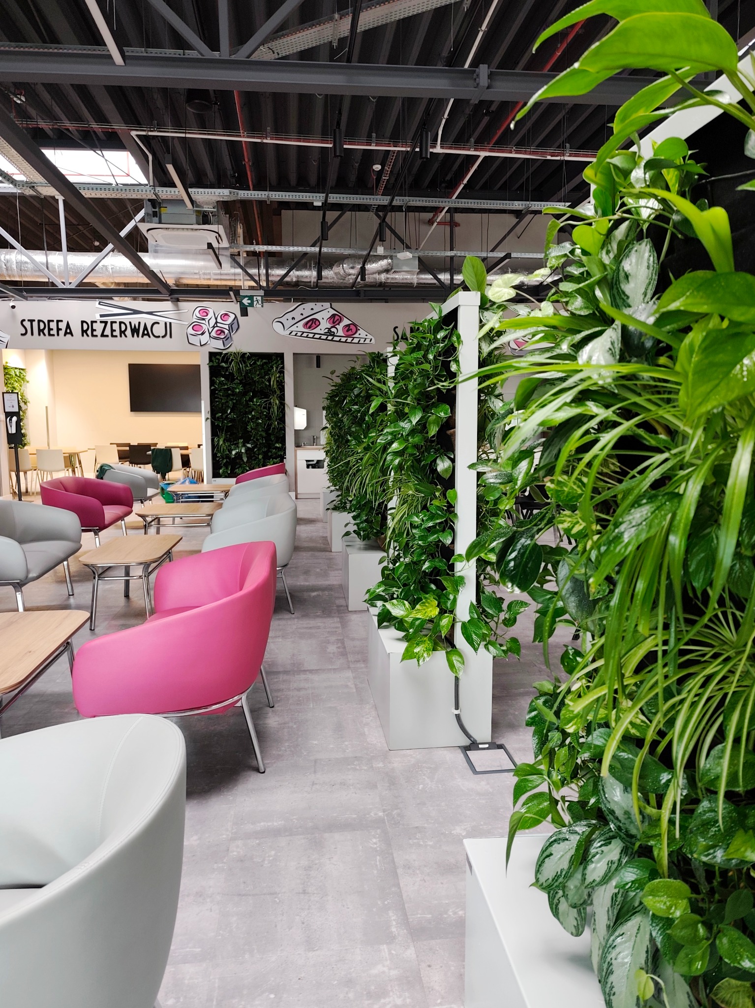 Wnętrze biurowe ze ścianami z roślin doniczkowych, strefa relaksu z kolorowymi fotelami i stolikami. Widoczne instalacje pod sufitem i napisy 'Strefa Rezerwacji'.