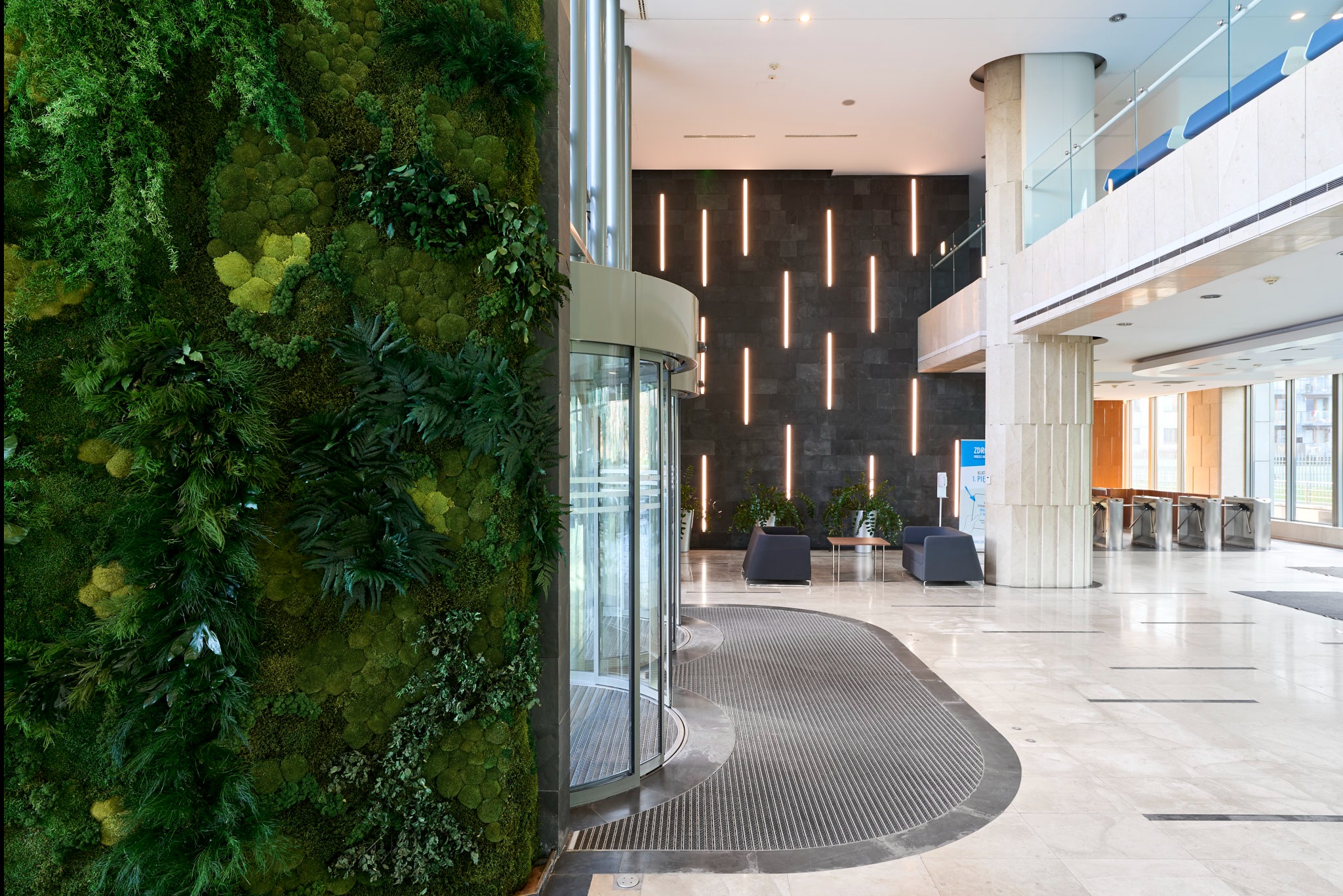 Wnętrze budynku z żywą ścianą z mchu i roślin, obok obrotowe drzwi wejściowe. Nowoczesny design z elementami natury.