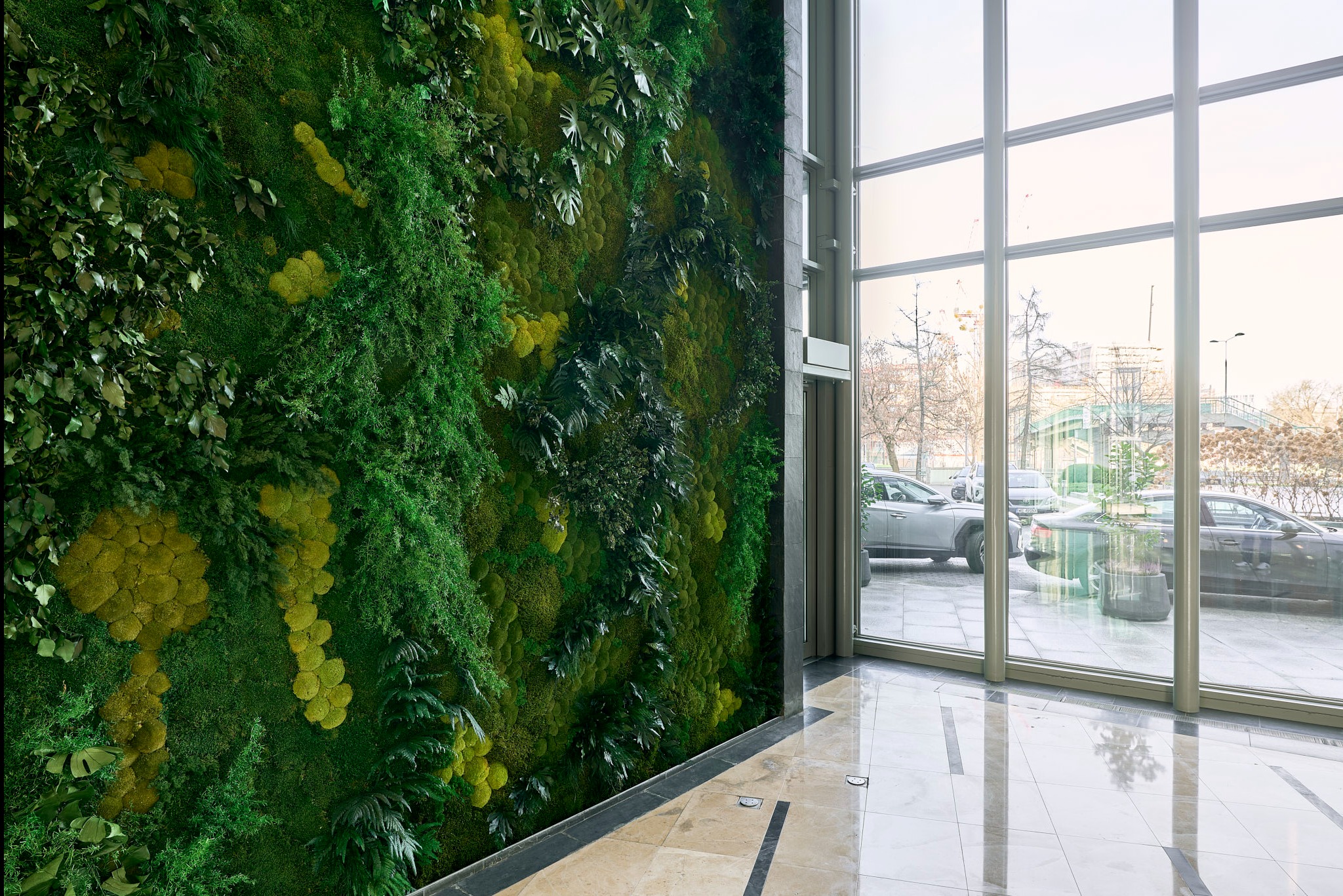 Wnętrze z pionową ścianą z roślin, widok na zewnątrz przez duże okna. Elementy zieleni w różnych odcieniach tworzą naturalny wzór na ścianie.