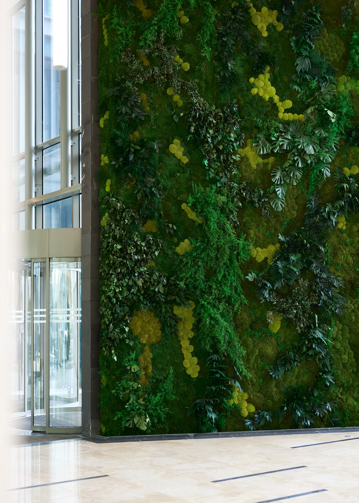 Żywa ściana z mchu i roślin w budynku biurowym, obok szklanych drzwi obrotowych i marmurowej podłogi. Dekoracja wnętrz z elementami natury.